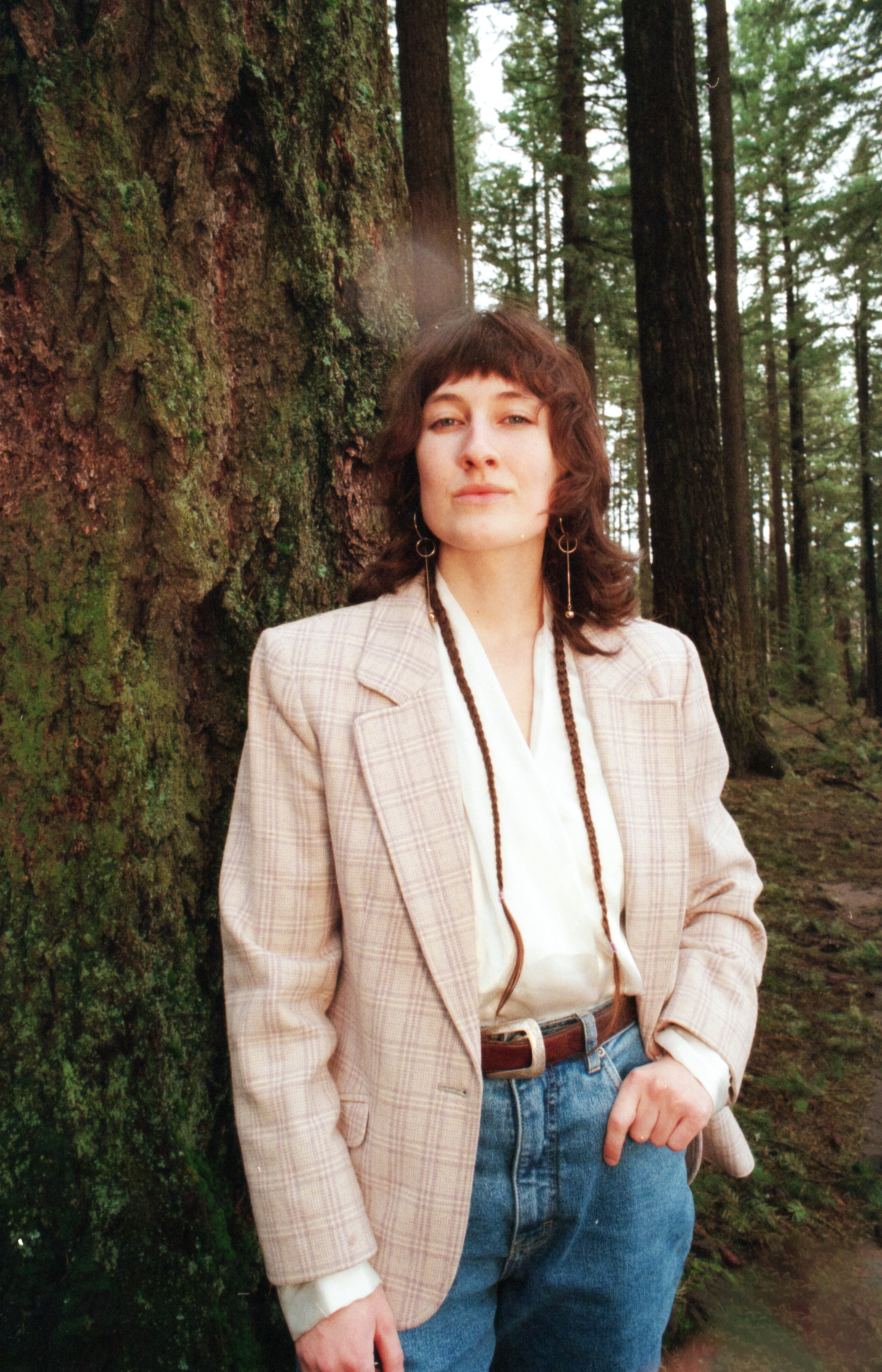 Hazelle Lerum stands in a forest wearing a plaid white/purple blazer, gold dangly earrings, and hair with two braids across her chest. She is in front of a tree, expression slightly smirking.